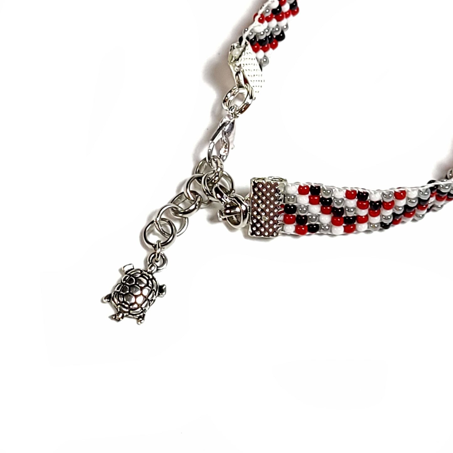 Clasp and charm close up of a woven handmade seed bead bracelet with red, white, black and light gray seed beads. Silver toned end clips with silver toned lobster clasp and silver toned turtle charm.