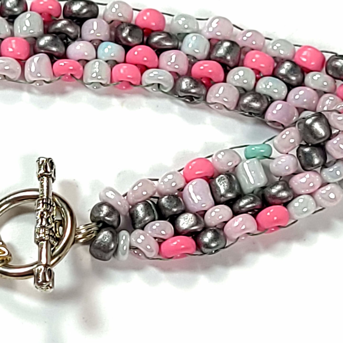 Close up of Bracelet made of irregular sized bright pink, white, pale pink, dark gray seed beads with one rebellious green bead. It has a toggle clasp.