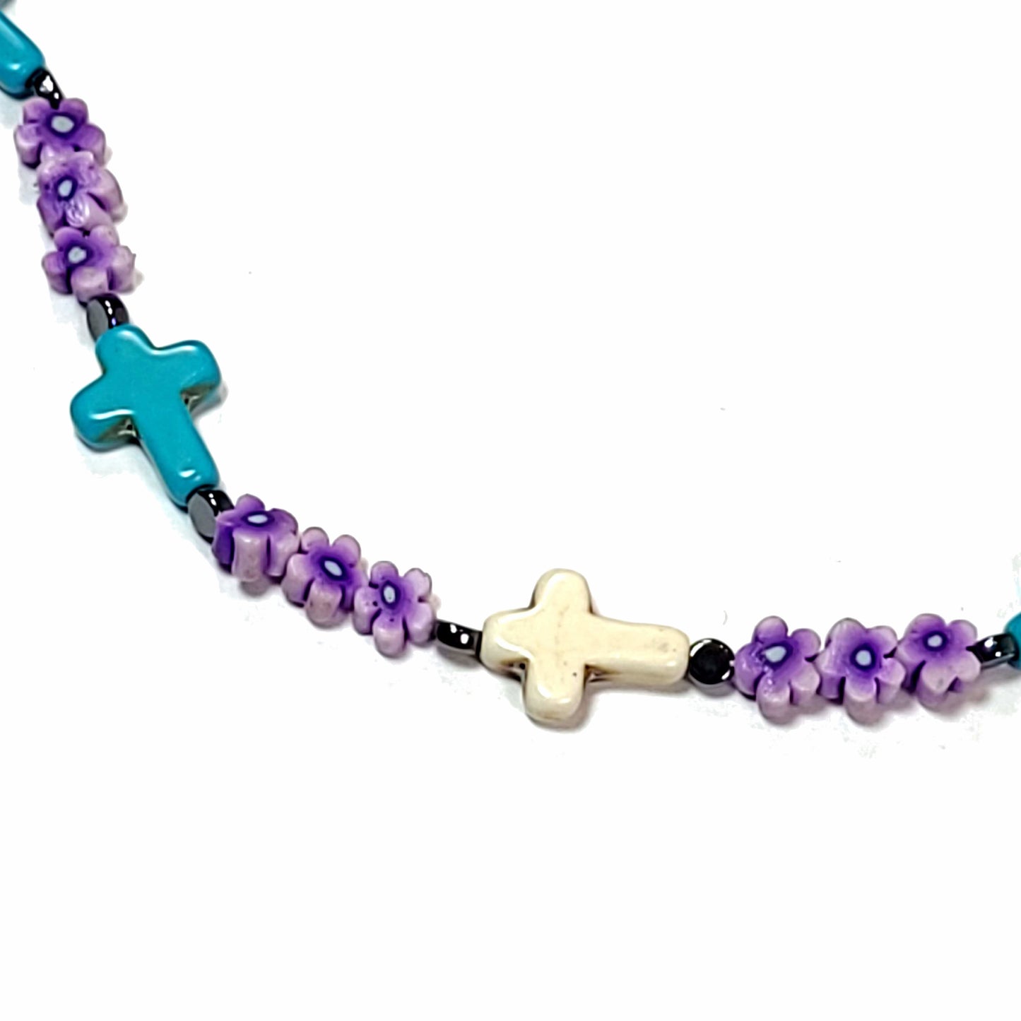 Close up of a polymer clay bead necklace with a blue cross, a white cross, small black beads. and purple floral beads.