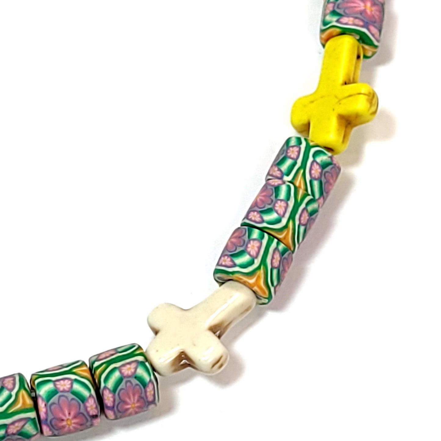 Close up of a Polymer bead necklace with cylindrical beads that have a floral pattern, and a yellow crosses, and a white cross.