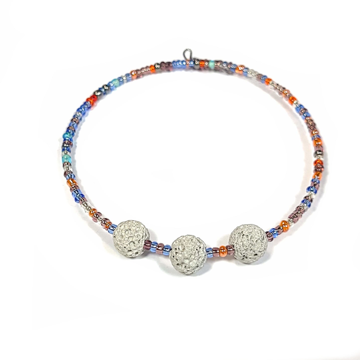 Bracelet and earring set made of multi-color seed beads in the blue and orange color range. The bracelets (2) are singular memory wire with triple lava rock center pieces. Small hoop earrings drop approximately. View of bracelet with white lava rocks.