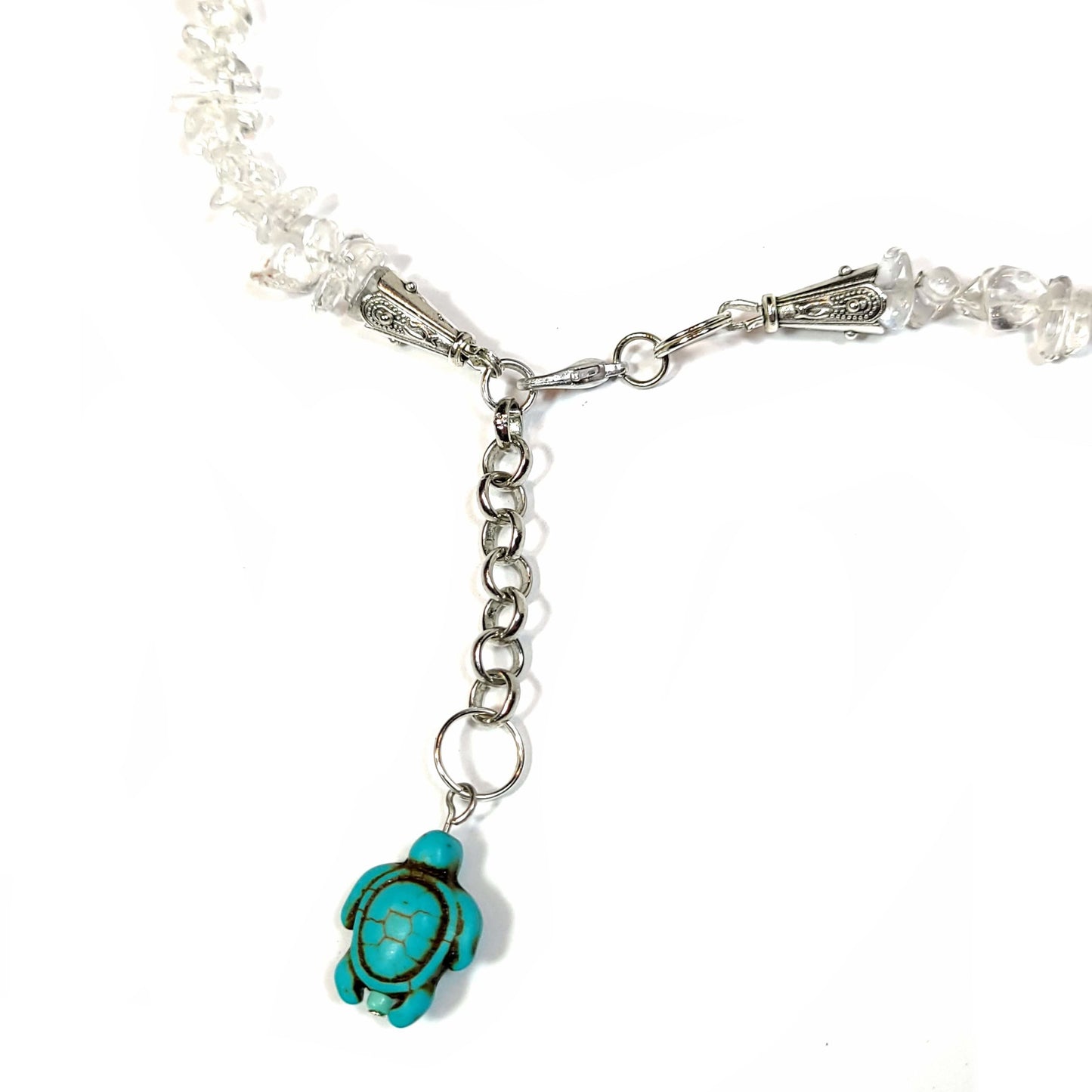Close up of the clasp and dangle turtle charm on a Necklace made of crystal quartz chips with aqua dyed howlite, with silver tone bale for center turtle and end caps. Silver tone extender chain and lobster clasp.
