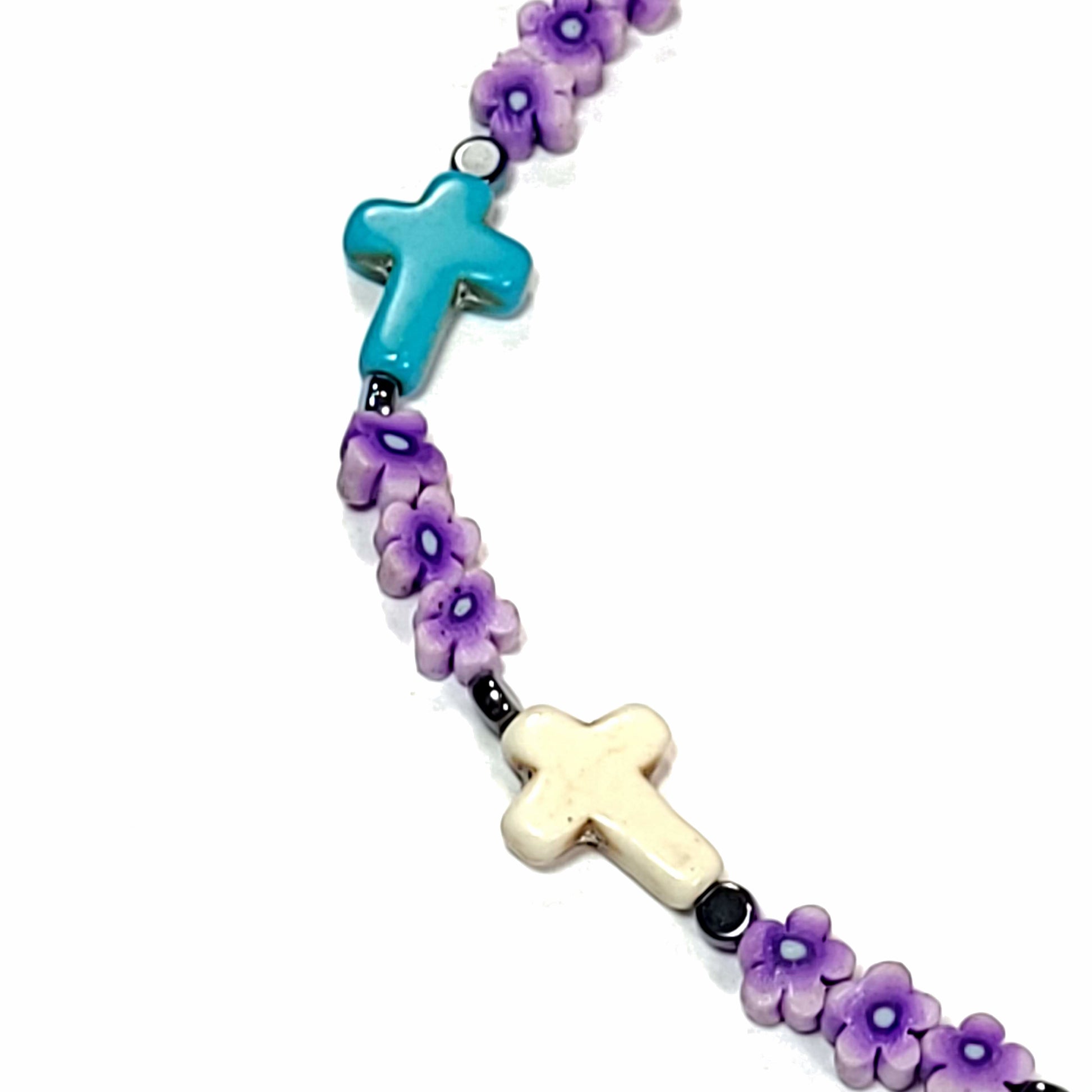 Close up of a polymer clay bead necklace with a blue cross, a white cross, small black beads. and purple floral beads.