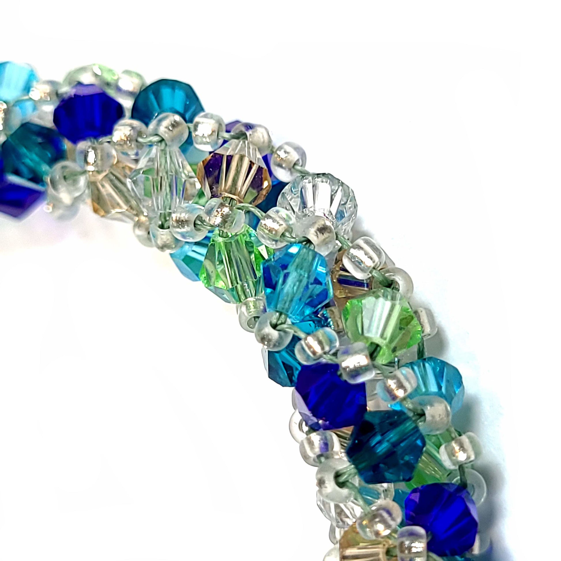 Close up view of a Bracelet made of teal, navy, light green, light blue, sand and crystal bicone beads with silver tone seed beads. It also has silver tone end caps, lobster clasp and extender chain. 